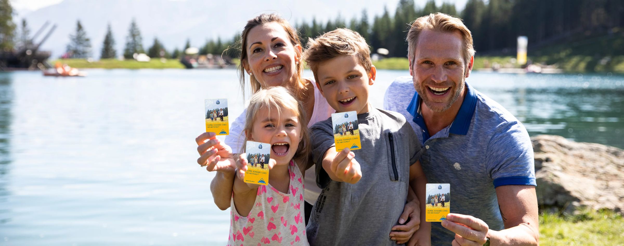 Lachende Familie hält am Bergsee die Super. Sommer. Card. in die Kamera in Serfaus-Fiss-Ladis