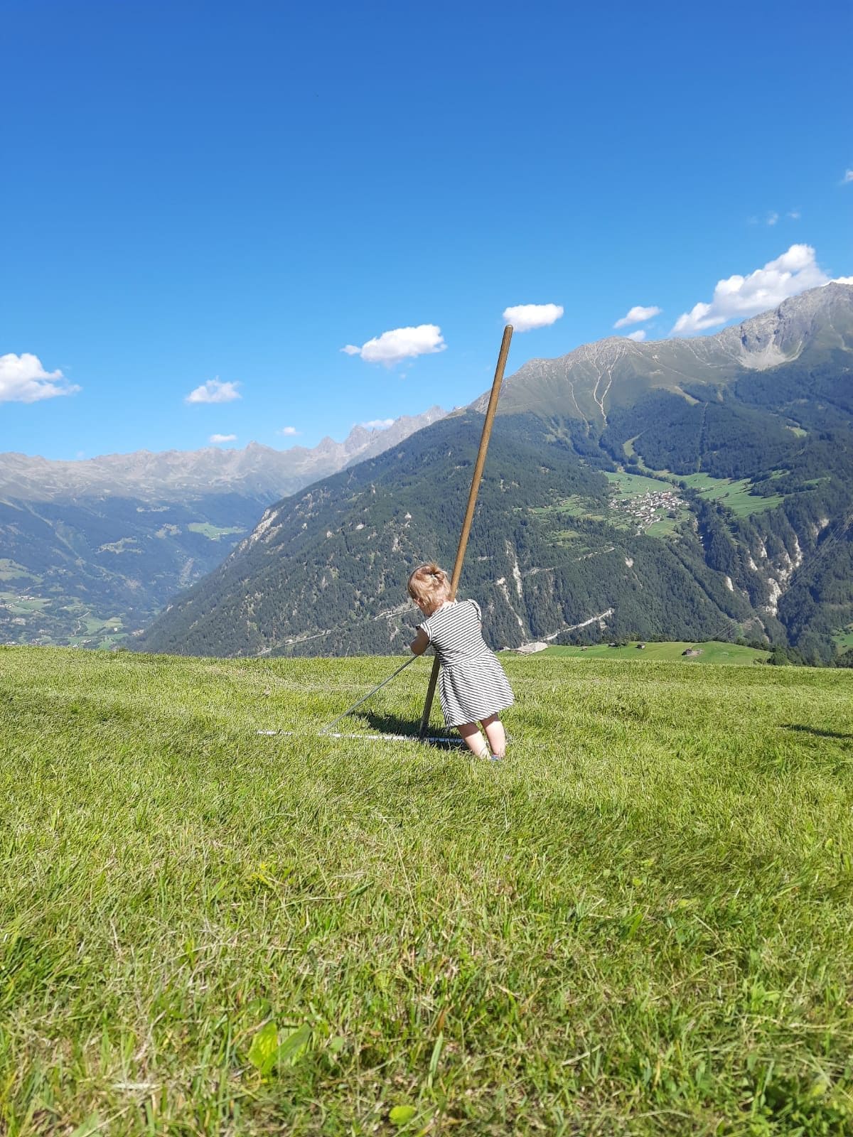 Kleines Kind mit Holzrechen auf einer sonnigen Bergwiese hoch über dem Tal am Sonnenplateau