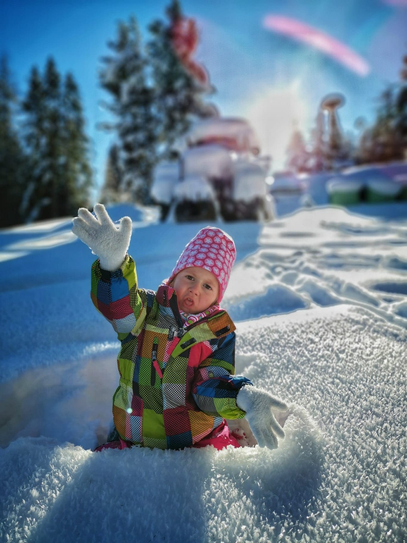 Kleinkind im bunten Schneeanzug winkt fröhlich im tiefen Neuschnee an einem sonnigen Wintertag