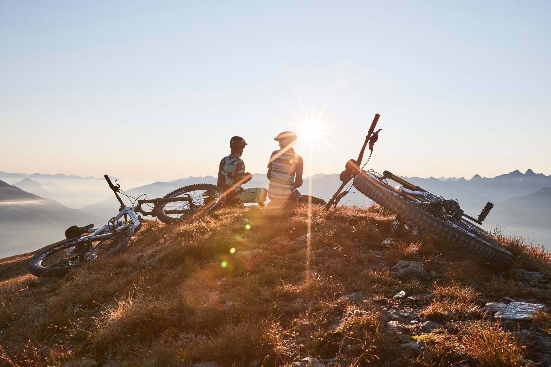 Zwei Mountainbiker rasten bei Sonnenuntergang auf einem Gipfel über dem Tal in Serfaus-Fiss-Ladis