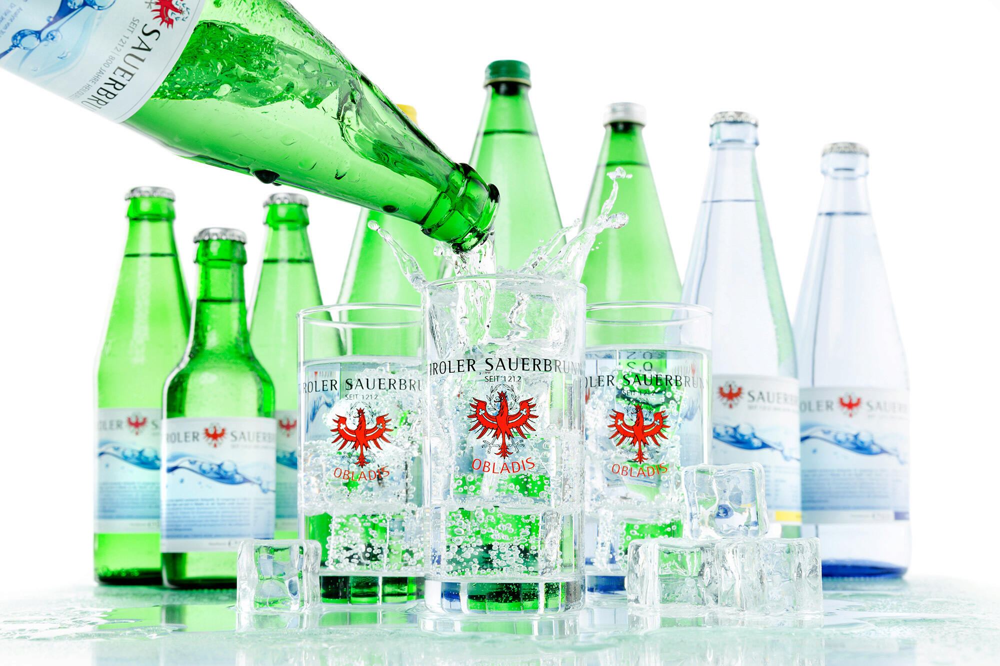 Eingeschenkter Tiroler Sauerbrunn in Gläsern mit Obladis-Logo, umgeben von grünen und blauen Flaschen und Eiswürfeln